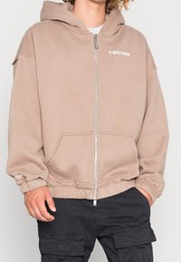 Sudadera con capucha beige con cremallera, cordón ajustable, bolsillos delanteros y puños acanalados. Presenta un logo impreso en el pecho y tela texturizada.