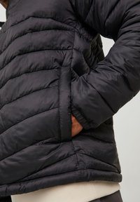 Svart boblevott med quiltet design, med høy krage og elastiske mansjetter. Stoffet har en glatt, glinsende tekstur.