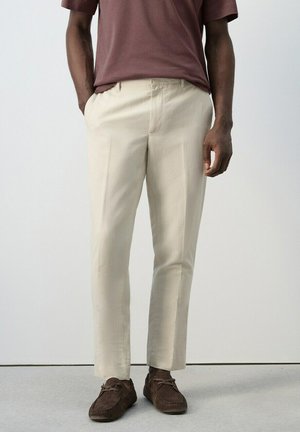 Hombre con pantalones beige de pierna recta, mocasines de ante marrón oscuro y camiseta de manga corta granate, con las manos en los bolsillos.