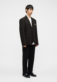 Άνδρας που στέκεται φορώντας ένα υπερμεγέθες μαύρο σακάκι, λευκό πουκάμισο, μαύρο παντελόνι και μαύρα παπούτσια επίσημα, μπροστά σε ένα απλό λευκό φόντο.