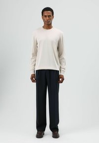 Beige shirt met lange mouwen en een ronde hals, gecombineerd met donkerblauwe broek en bruine schoenen, allemaal van effen stof zonder patronen.