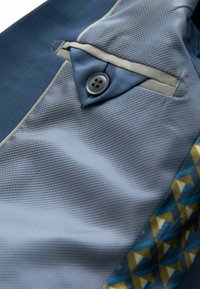 Un tessuto blu texturizzato presenta una piccola tasca con un bottone grigio e un motivo geometrico in blu e oro lungo il bordo.