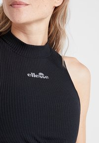 Femme portant un haut sans manches noir côtelé avec un col montant et le logo « ellesse » imprimé sur la poitrine.
