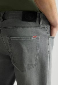 Los jeans de mezclilla gris presentan un diseño ajustado, un parche de cuero en la cinturilla y una pequeña etiqueta de logo rojo en el bolsillo trasero.