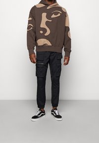 Uomo che indossa un felpa marrone con design di volti astratti, pantaloni cargo neri con numerose tasche e sneakers in bianco e nero.