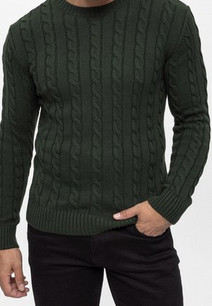 Man wearing dark green cable-knit sweater and black pants, with one hand in pocket against plain background.