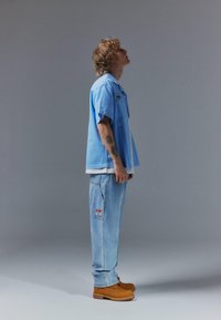 Jeune homme aux cheveux bouclés portant une chemise à manches courtes bleu clair, un jean ample avec un patch, des bottes marron, debout de profil devant un fond gris.