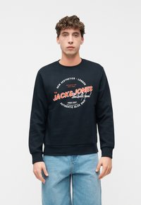 Νέος άνδρας που φοράει μαύρο φούτερ Jack & Jones με κόκκινο και λευκό κείμενο, συνδυασμένο με ανοιχτό μπλε τζιν, στέκεται μπροστά σε απλό φόντο.