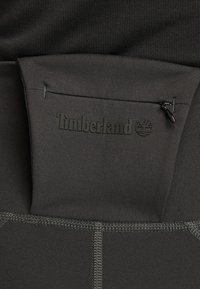 Svarta atletiska shorts har en åtsittande midja med en dragkedjeficka. Tyget verkar slätt med subtila sömdetaljer och en Timberland-logotyp.