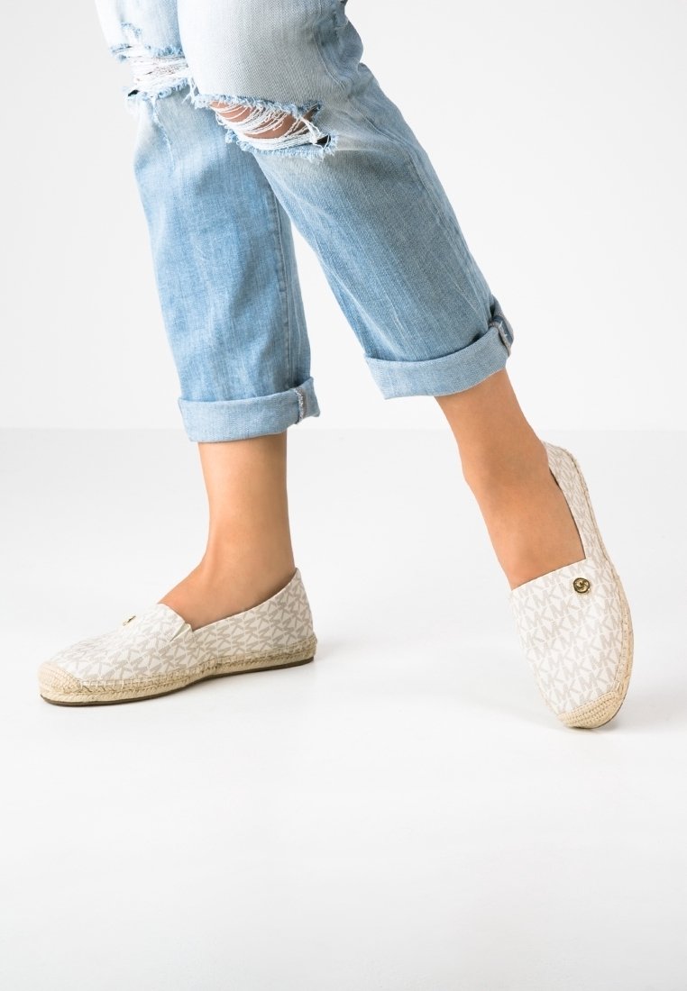 Pieds portant des chaussures slip-on blanches à motifs, associées à un jean bleu clair déchiré et roulé sur un fond blanc uni.