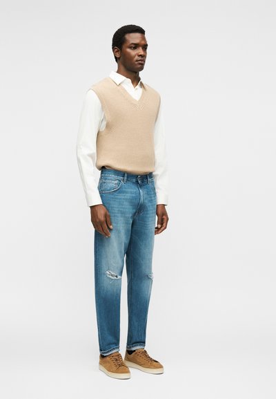 Άνδρας που φοράει μπεζ πλεκτό γιλέκο πάνω από λευκό πουκάμισο, σκισμένα μπλε τζιν και καφέ σουέντ αθλητικά παπούτσια, στέκεται μπροστά σε απλό φόντο.