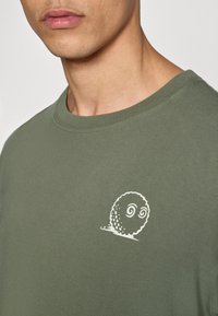 Πράσινο ελιάς βαμβακερό t-shirt με στρογγυλή λαιμόκοψη, με λευκό γραφικό απεικόνισης ενός καρτούν σαλιγκαριού στο αριστερό στήθος. Ομαλή υφή.