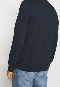 Granatowa bluza z długimi rękawami i okrągłym dekoltem, o gładkiej fakturze i ściągaczowych wykończeniach, zestawiona z jasnoniebieskimi jeansami.