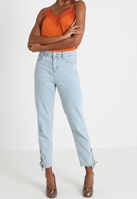 Orange camiscule med spetsdetaljer, ljusblå högmidjade jeans med fransar och sidoklipp, tillsammans med mönstrade högklackade skor.