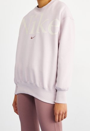 Personne portant un sweat Nike rose clair et des leggings mauves, debout devant un fond clair uni.