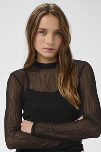 Jauna moteris su ilgais rudais plaukais, dėvinti juodą permatomą ilgomis rankovėmis megztinį ant juodos kamizolės, sukryžiavusi rankas, neutralus veido išraiška.