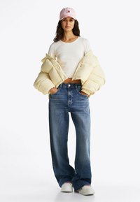 Giacca puffer gialla chiara, top bianco a maniche lunghe, jeans blu larghi con scintillii e sneaker bianche. Cappellino rosa con dettagli del logo.