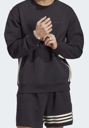 Person mit schwarzem Sweatshirt und Shorts mit weißen Streifen, die Hände gefaltet, trägt Armbänder und Ringe, von den Schultern bis zu den Oberschenkeln gezeigt.