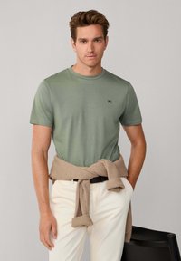 T-shirt verde oliva a maniche corte in cotone, con un piccolo logo blu navy sul petto, abbinata a pantaloni color crema e a un maglione beige legato intorno alla vita.