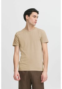 Beige, rövid ujjú póló puha anyagból, kerek nyakkivágással és testhez simuló fazonnal. Barna, strukturált nadrággal viselve.