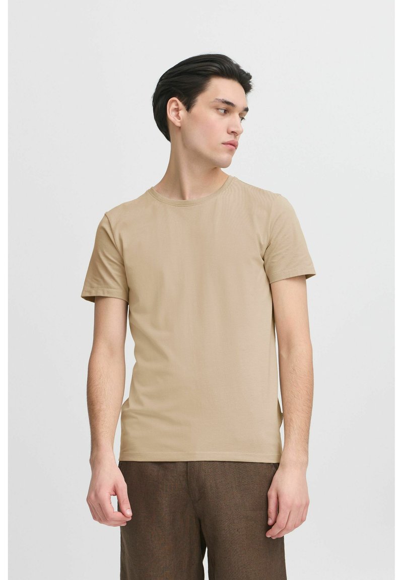 Beige, rövid ujjú póló puha anyagból, kerek nyakkivágással és testhez simuló fazonnal. Barna, strukturált nadrággal viselve.