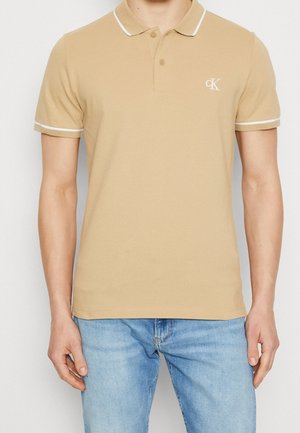 Tan polo shirt med hvide detaljer på kraven og ærmerne. Har en tre-knaps knappelukning og broderet logo på brystet. Lavet af en bomuldsblanding.