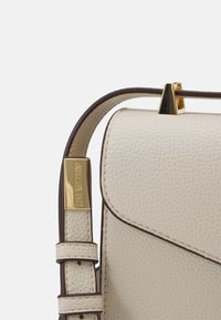 Borsa a tracolla in pelle color crema con superficie testurizzata, hardware in tono oro e una tracolla in accento marrone scuro. Presenta una chiusura a pattina.