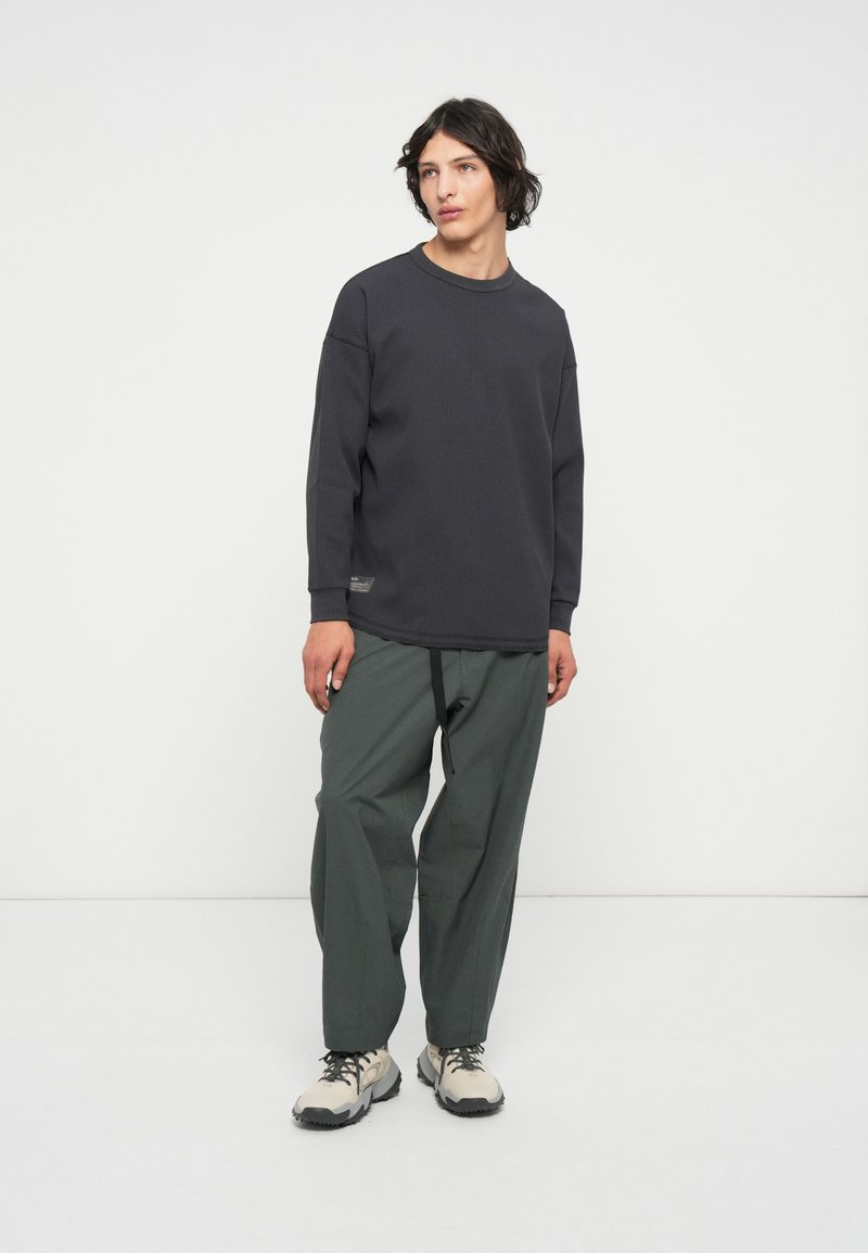 Chemise à manches longues texturée grise associée à un pantalon vert ample et des baskets beiges, avec une poche latérale et des coutures contrastées.