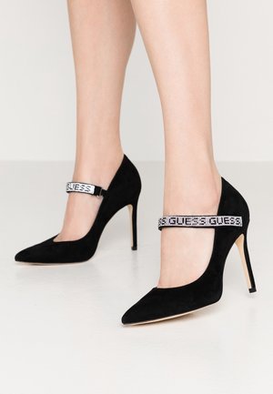 Escarpins à talons hauts en daim noir avec bouts pointus et brides ornées de strass affichant le nom de la marque "GUESS", portés pieds nus.