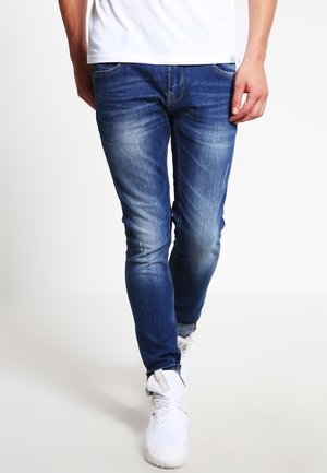 Jambes d’une personne portant un jean bleu slim avec des détails délavés, des baskets blanches et un t-shirt blanc sur un fond clair uni.