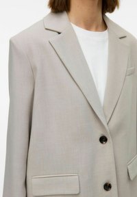 Vaalea harmaa bleiseri, jossa on leikkausläppä, kaksi nappia ja vasemmalla puolella rintatasku. Siinä on sileä pinta ja tyylikäs istuvuus.