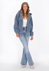 Donna sorridente, con indosso un felpa blu con zip, una canotta bianca, jeans chiari a zampa d'elefante e scarpe beige a punta, che sta in piedi davanti a uno sfondo bianco.