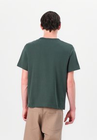 T-shirt en coton vert foncé avec un col rond et des manches courtes, présentant une coupe droite et une texture lisse ; porté avec un pantalon beige rayé.
