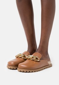 Mocassins bruns à enfiler avec un grand détail de chaîne dorée, portés sur des pieds à la peau foncée sur fond blanc.