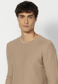 Beige bordázott, hosszú ujjú pulóver kerek nyakkal és karcsú fazonnal. Az anyag textúrája enyhén kiemelkedő, szemet gyönyörködtető mintát adva.