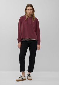 Mujer de pie con sudadera burdeos, pantalones cortos negros, camiseta a rayas debajo y zapatillas beige con detalles negros en un fondo liso.