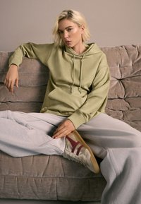 Zelená mikina s kapucňou a sťahovacími šnúrkami, v kombinácii so sivými teplákmi. Béžové tenisky s červenými detailmi. Modelka sediaca na textúrovanom béžovom pohovke.