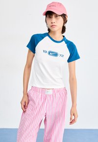 Bela kratka majica s kratkimi rokavi in modrimi raglan rokavi, na kateri je logo "Nike 1972"; v kombinaciji z ohlapnimi roza in belimi črtastimi hlačami.