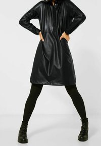 Svart kjol i konstläder med knäppning framtill, långa ärmar och sidofickor. Har en lös passform och en mjuk textur.