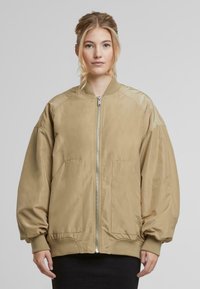Khaki värvi bomber-jakk, mis on valmistatud siledast kangast, millel on ees lukuga sulgur, kaks külge taskut, ribakudumine mansettides ja lõdvestatud istuvus.