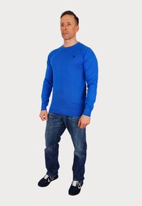 Modra pletena pulover s krožnim izrezom, oblečen over modrih kavbojkah. Ima majhen temen logotip na levi prsni strani. Kopačke dopolnijo obleko.