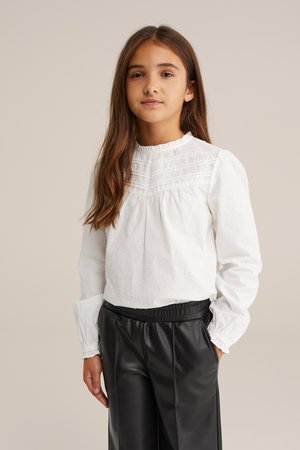 Blusa blanca de manga larga con una textura sutil y pequeños puntos, que presenta un escote con volantes y un cuerpo fruncido, acompañada de pantalones de imitación cuero negros.