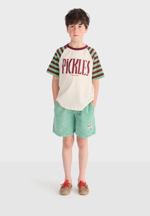 Jongen staat met handen in de zakken, draagt een wit en groen gestreept T-shirt met "PICKLES" erop, groene korte broek, en bruine schoenen met rode veters.