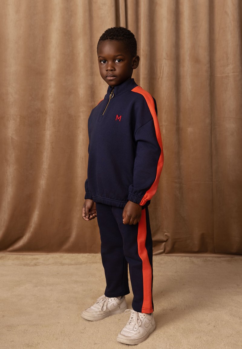 Mornarska trenirka z rdečimi stranskimi črtami, z zadrgo na ovratniku in izvezenim logotipom 'M'. Obuta z belimi supergami. Mehka tekstura, vsakdanji dizajn.