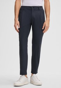 Granatowe spodnie z teksturowanej tkaniny w slim fit, z zapięciem na guzik, elastycznym pasem i przednimi kieszeniami. W zestawieniu z białymi butami.