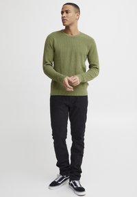 Grüner, gestrickter Rundhalspulli, taillierte Form, mit geripptem Muster. Kombiniert mit schwarzen Hosen und schwarzen Sneakers mit weißen Akzenten.