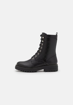 Botte de combat en cuir noir à lacets avec semelle épaisse à crampons et languette à l’arrière, montrée de côté extérieur sur un fond uni.