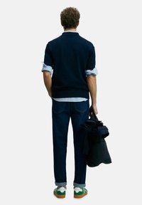 Mann, der mit dem Rücken zur Kamera steht, trägt einen marineblauen Pullover, hochgekrempelte gestreifte Hemdsärmel, umgeschlagene Jeans und grüne Sneakers. Er hält eine schwarze Jacke in der rechten Hand.