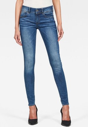 Vaqueros skinny de mezclilla, con una cintura de altura media, bolsillos frontales y un lavado desgastado. El modelo lleva puestos tacones altos negros.