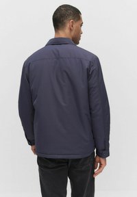 Marinblå jacka med kragdesign, knappade manschetter och en mjuk textur, visad bakifrån mot en enfärgad bakgrund.